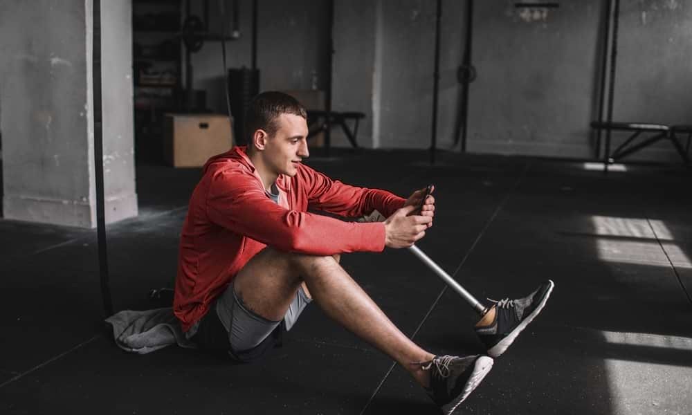 White man with prosthetic left leg sitting on gym floor, using smartphone