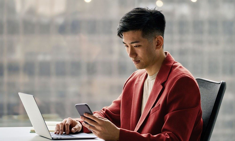 Asian young adult man sitting at laptop with smartphone in hand